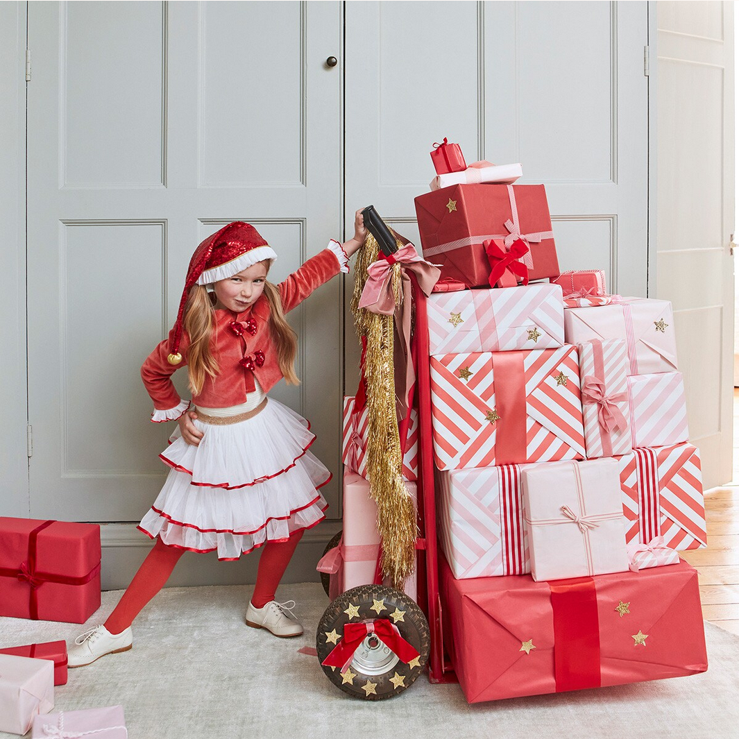 Child in festive outfit standing next to a stack of wrapped gifts in a room.