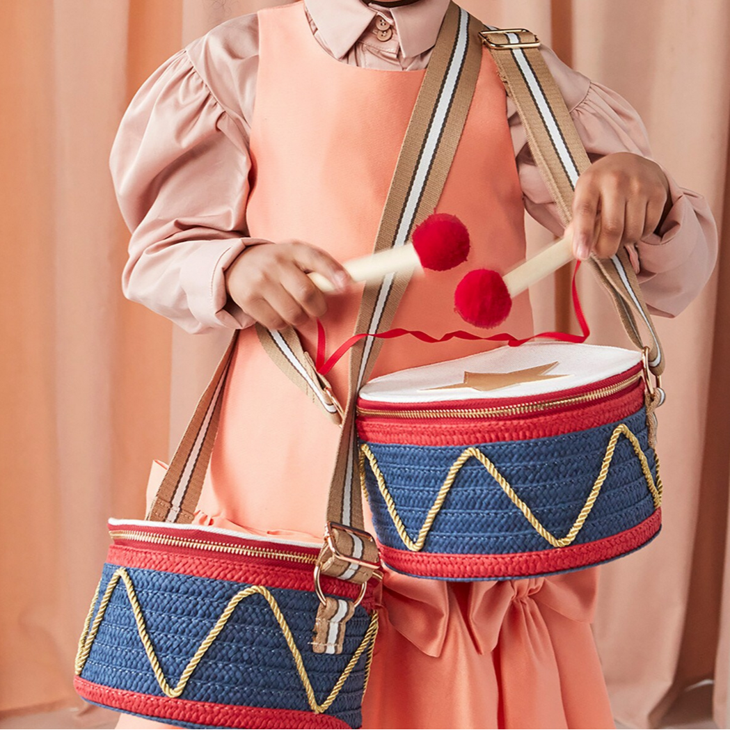 Person playing a small drum with red pom-poms against a peach-colored curtain background