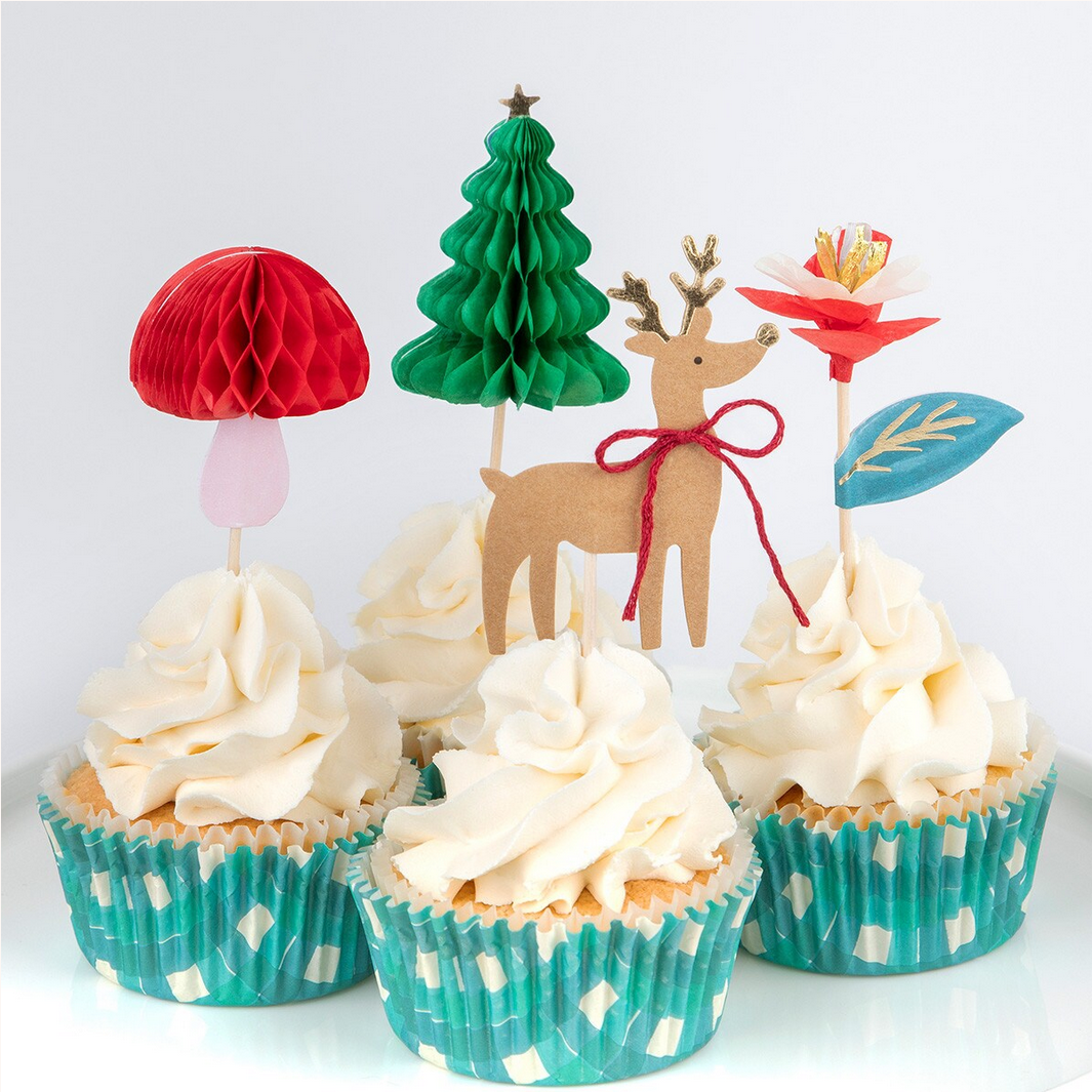 Cupcakes with decorative toppers including a mushroom, tree, deer, and flower on a white background.