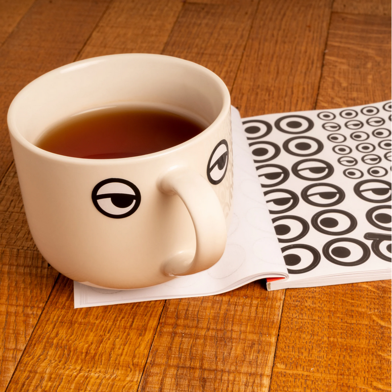 White mug with eye stickers filled with tea on a wooden surface next to a book with a black and white eye pattern.