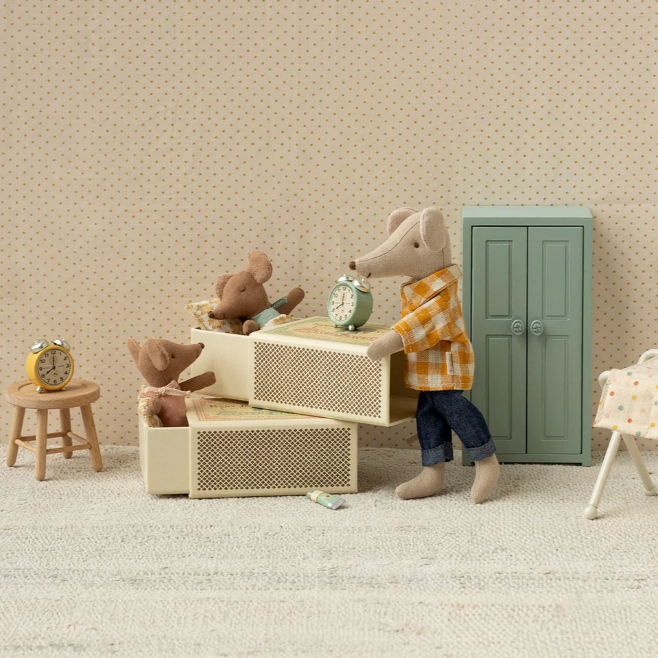 Small toy mouse in a plaid shirt standing next to a toy bed and dresser in a room with a patterned wall.