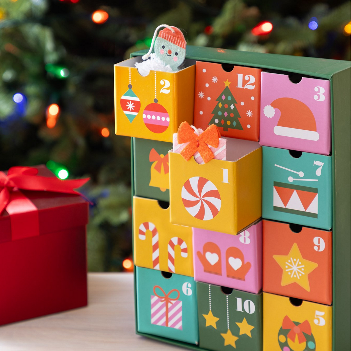 Colorful advent calendar with Christmas-themed designs in front of a decorated tree.