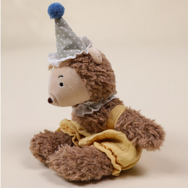 Plush toy hedgehog wearing a party hat and holding a yellow bag on a beige background