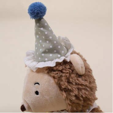 Plush hedgehog toy wearing a party hat with a blue pom-pom on a beige background