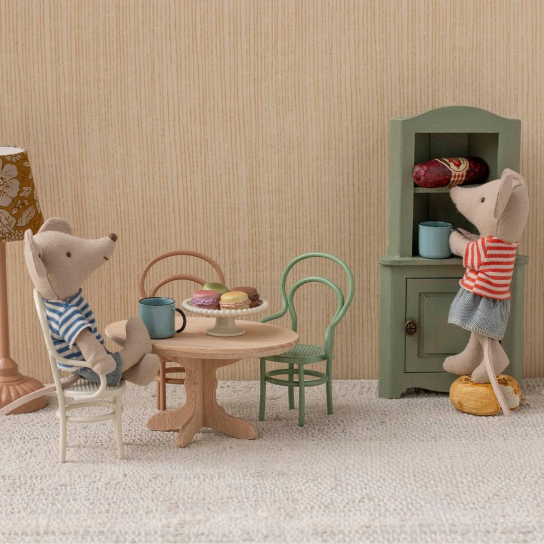 Toy scene with two teddy bears around a small table, lamp, and cabinet against a striped wall.