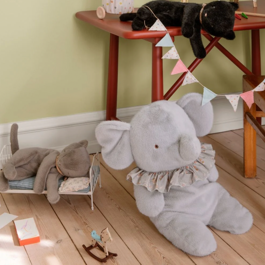 Gray elephant plush toy on a wooden floor with a child's play area in the background.