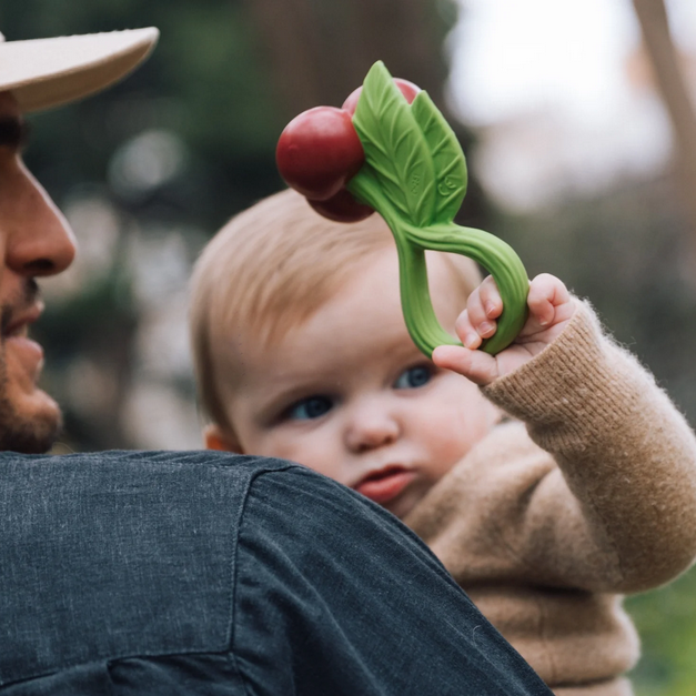 Baby holding a green and red teething toy with a blurred background