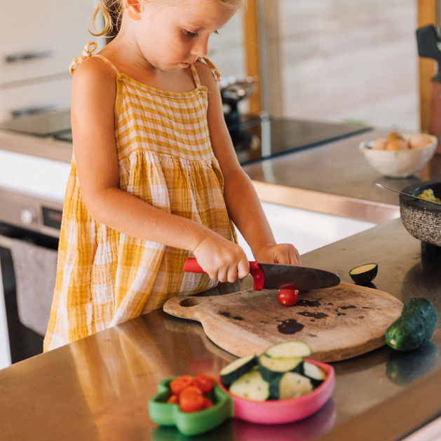 Child in a yellow dress cutting vegetables on a wooden board in a kitchen.
