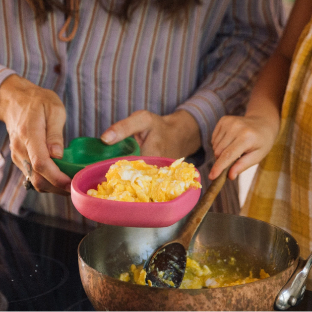 Person adding scrambled eggs from a pink bowl into a large metal pan.