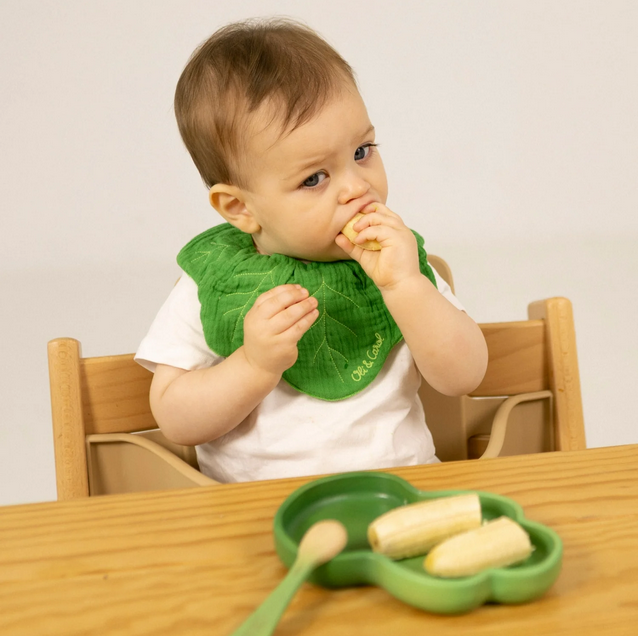 Baby wearing a green bib eating at a wooden table with a plain background