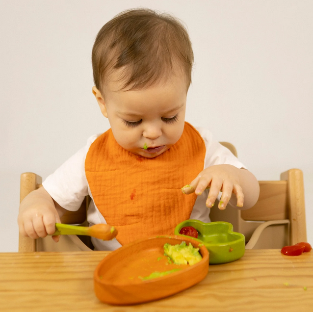 Baby in an orange bib eating with a spoon and bowl on a wooden table.