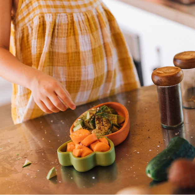 Child's hand reaching towards a bowl of food on a wooden table with a checkered dress.