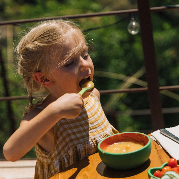 Child eating from a spoon outdoors with a bowl of soup on a table.