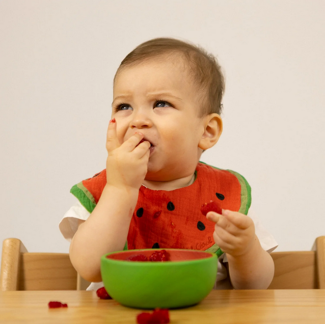 Baby wearing a watermelon-themed bib eating from a green bowl.