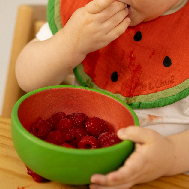 Child wearing a watermelon-themed bib eating raspberries from a green bowl.