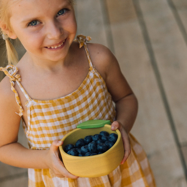 Young girl holding a yellow bowl of blueberries on a wooden floor.
