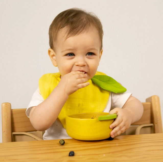Child wearing a yellow bib with a green leaf, sitting at a table with a bowl.