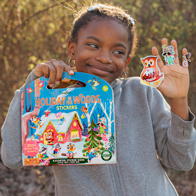 Child holding a box of 'Holiday Woods' stickers with owl stickers on fingers outdoors.