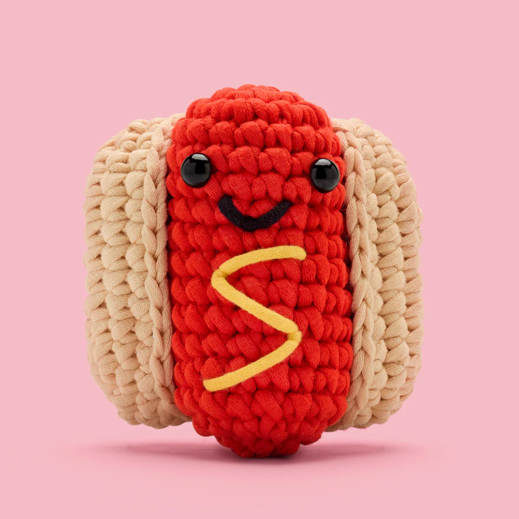 Crocheted toy resembling a hot dog with a face on a pink background