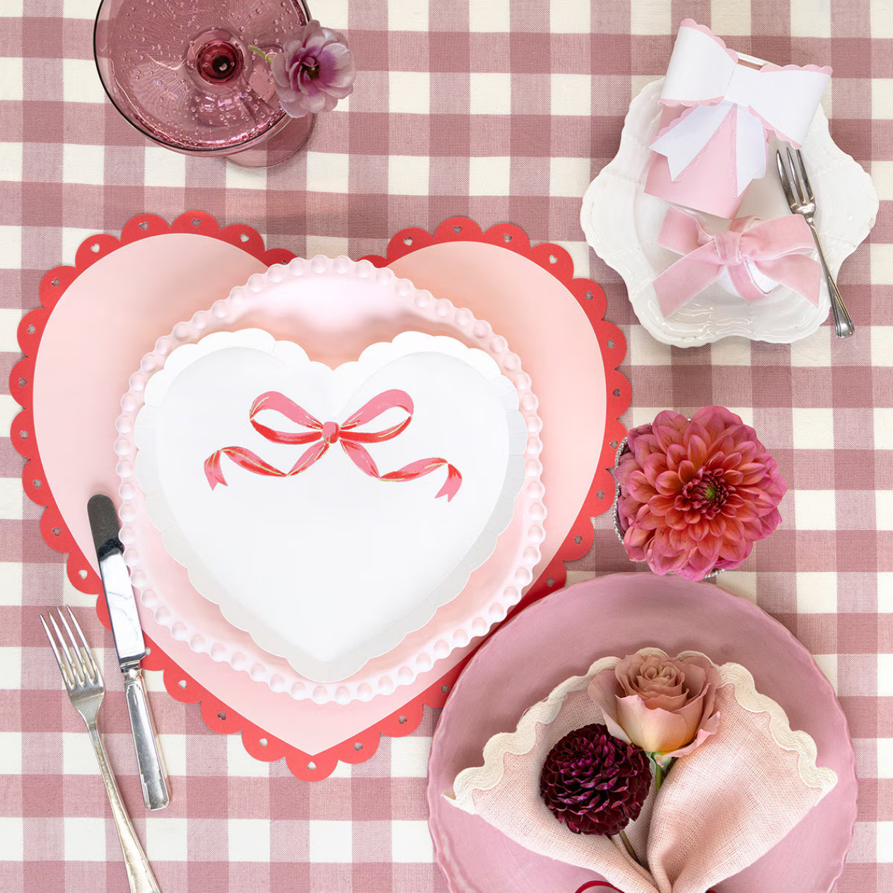 Heart-shaped cake with pink ribbon on a checkered tablecloth with floral and decorative elements.