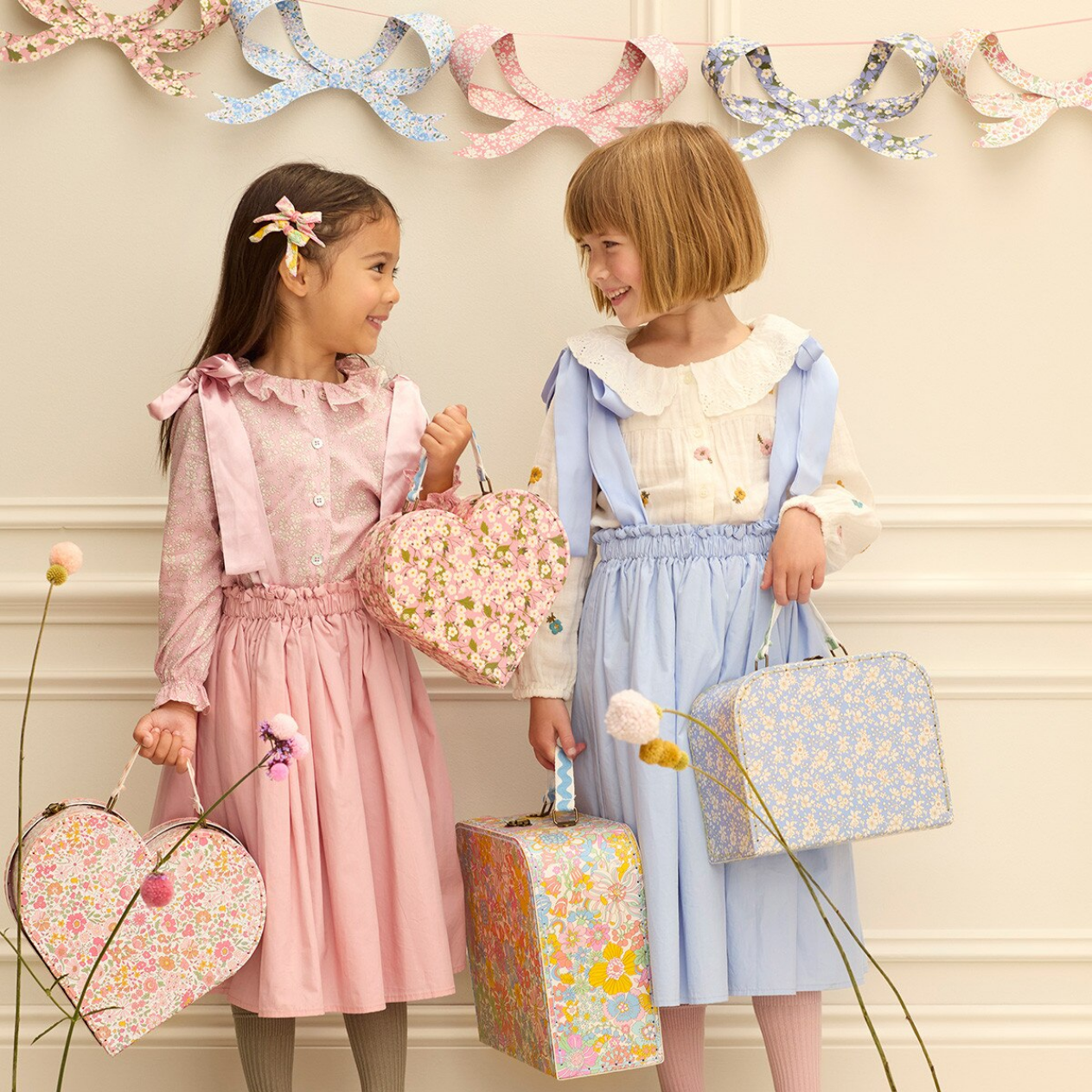 Two young girls in pastel dresses holding floral-patterned bags against a decorative wall.
