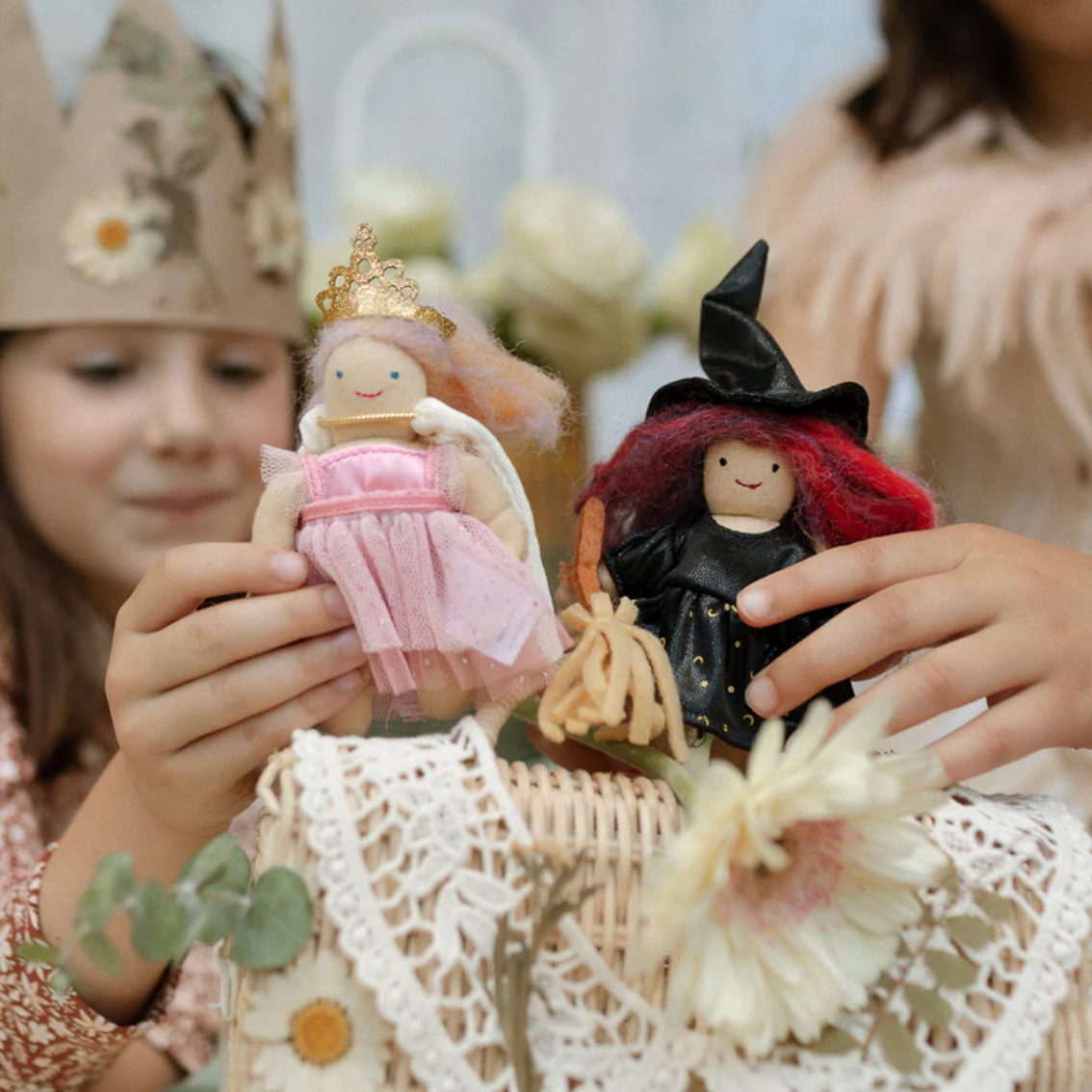Two children holding small dolls, one in a pink dress and crown, the other in a black witch outfit with a hat, against a blurred background.