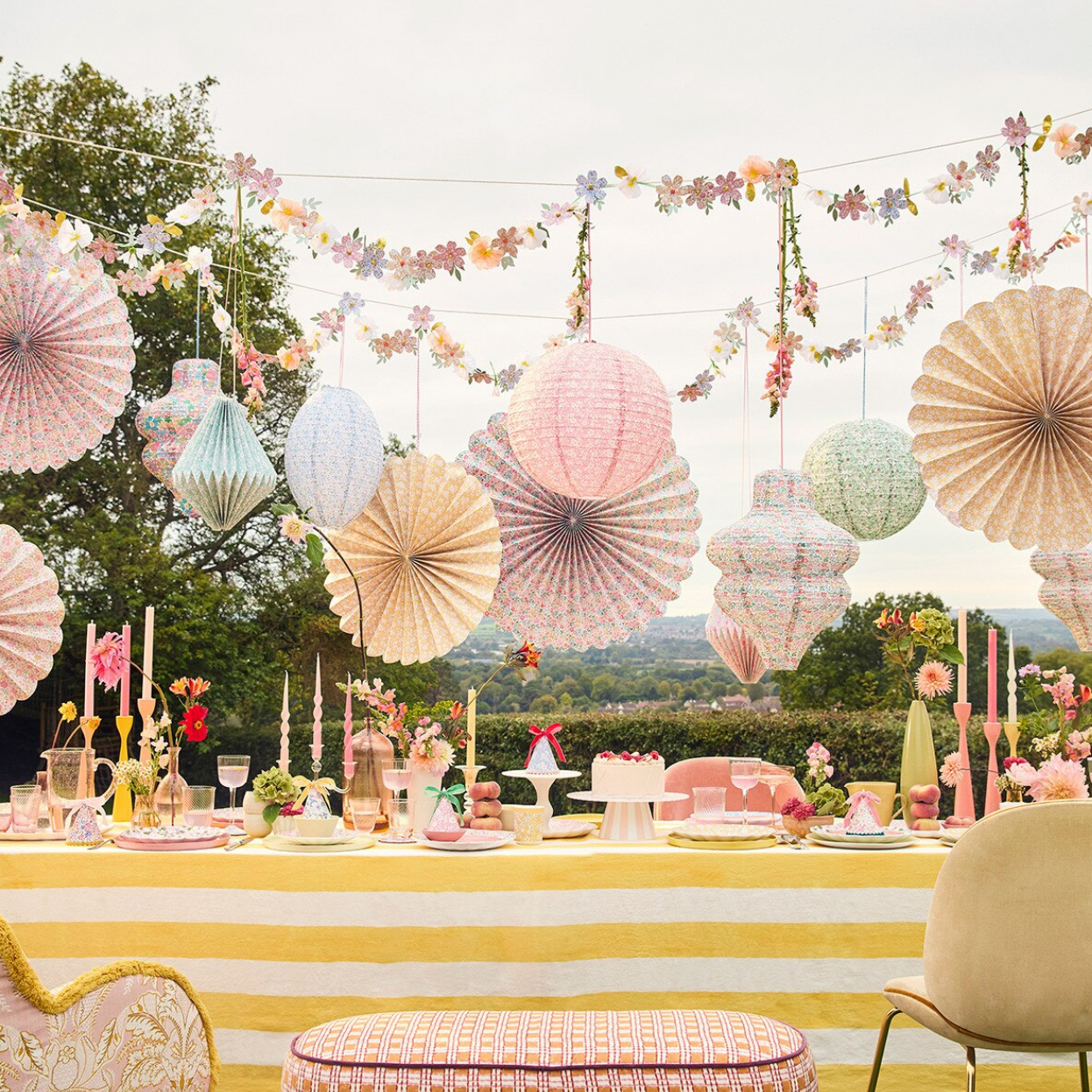 Decorative outdoor setting with hanging paper fans, floral garlands, and a table with candles and flowers.