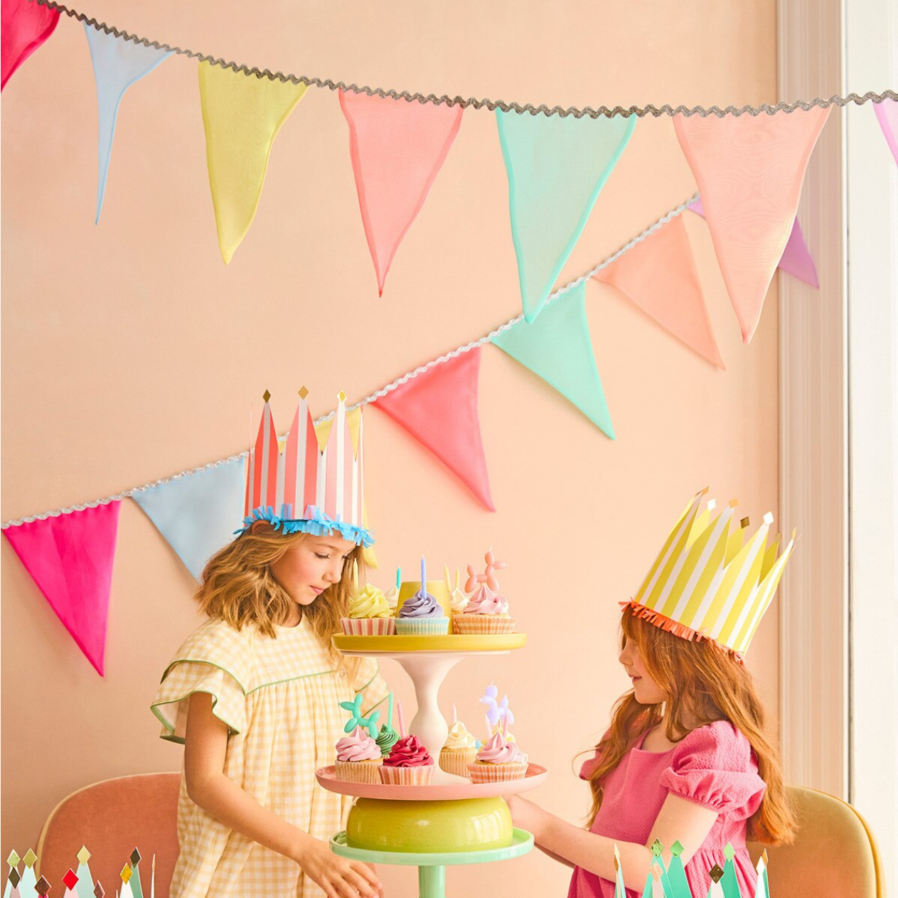 Two children wearing colorful birthday hats standing behind a tiered cake stand with small cakes, under a string of multicolored triangular flags.
