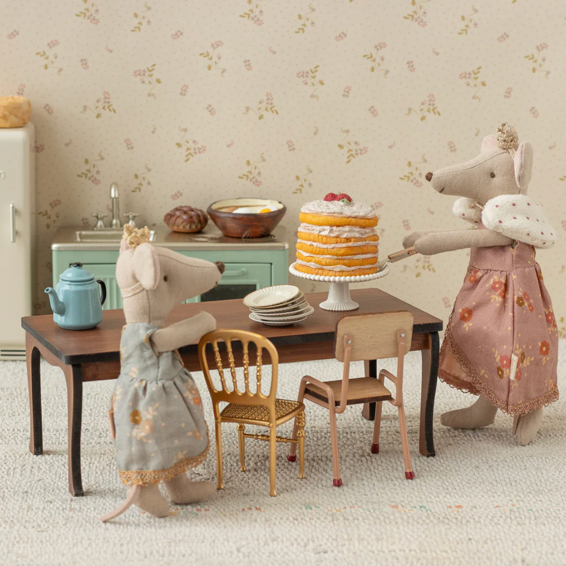 Two toy mice in dresses standing at a table with a cake in a room with floral wallpaper.
