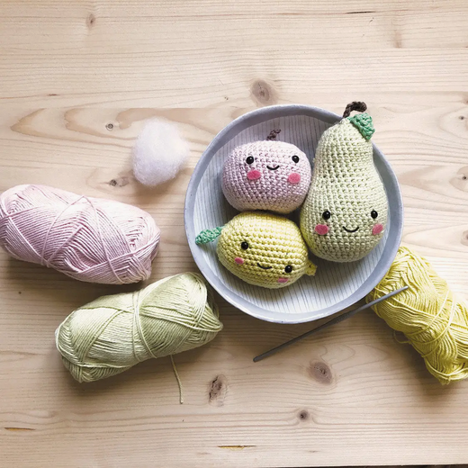Three crocheted pear-shaped toys with smiling faces in a bowl, surrounded by yarn and a crochet hook on a wooden surface.
