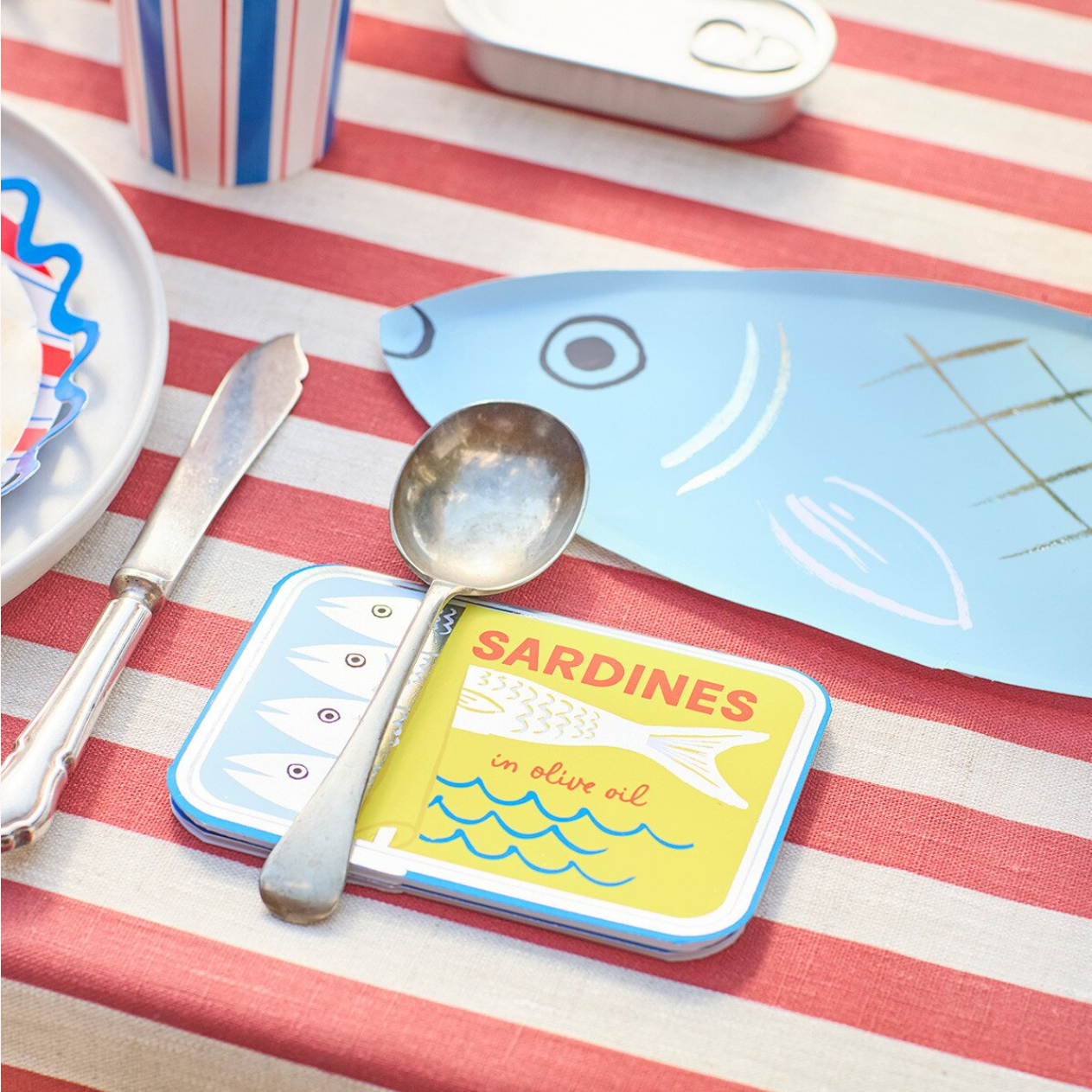 Sardine can with a spoon on a striped tablecloth with plates and cutlery.