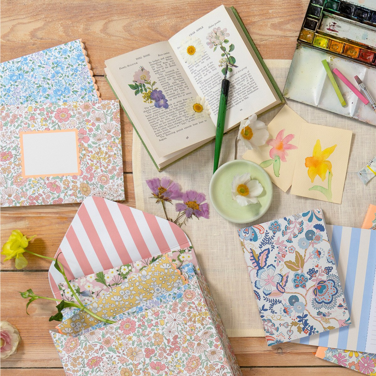 Floral stationery items including notebooks, cards, and a watercolor set on a wooden surface.