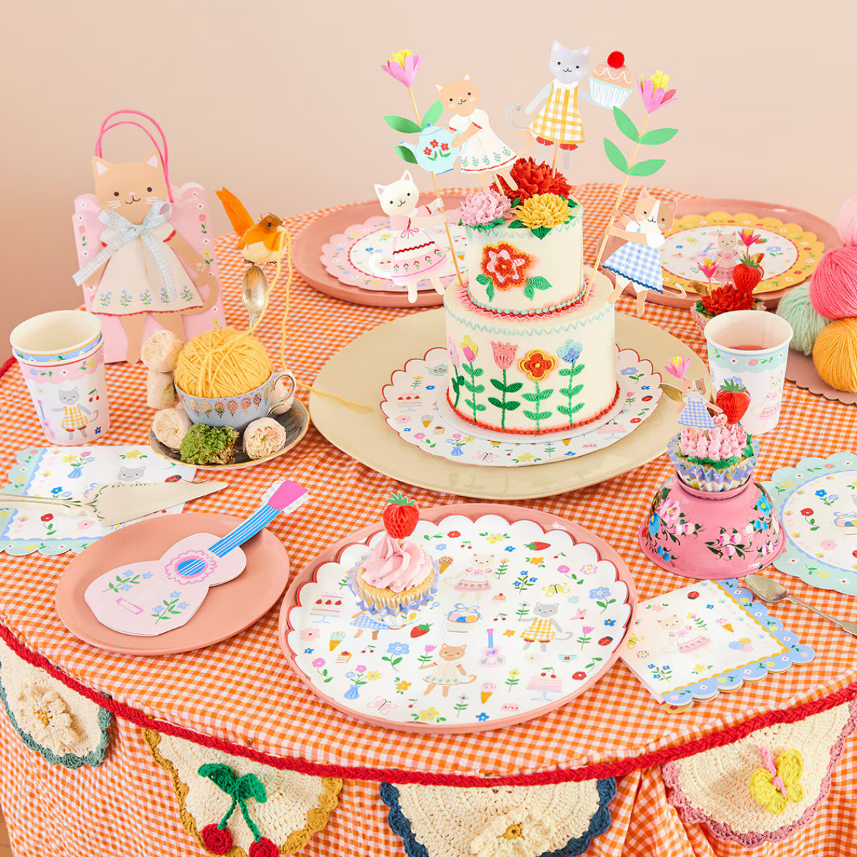 Children's play table with toy cake, dolls, and decorative items on a checkered tablecloth.