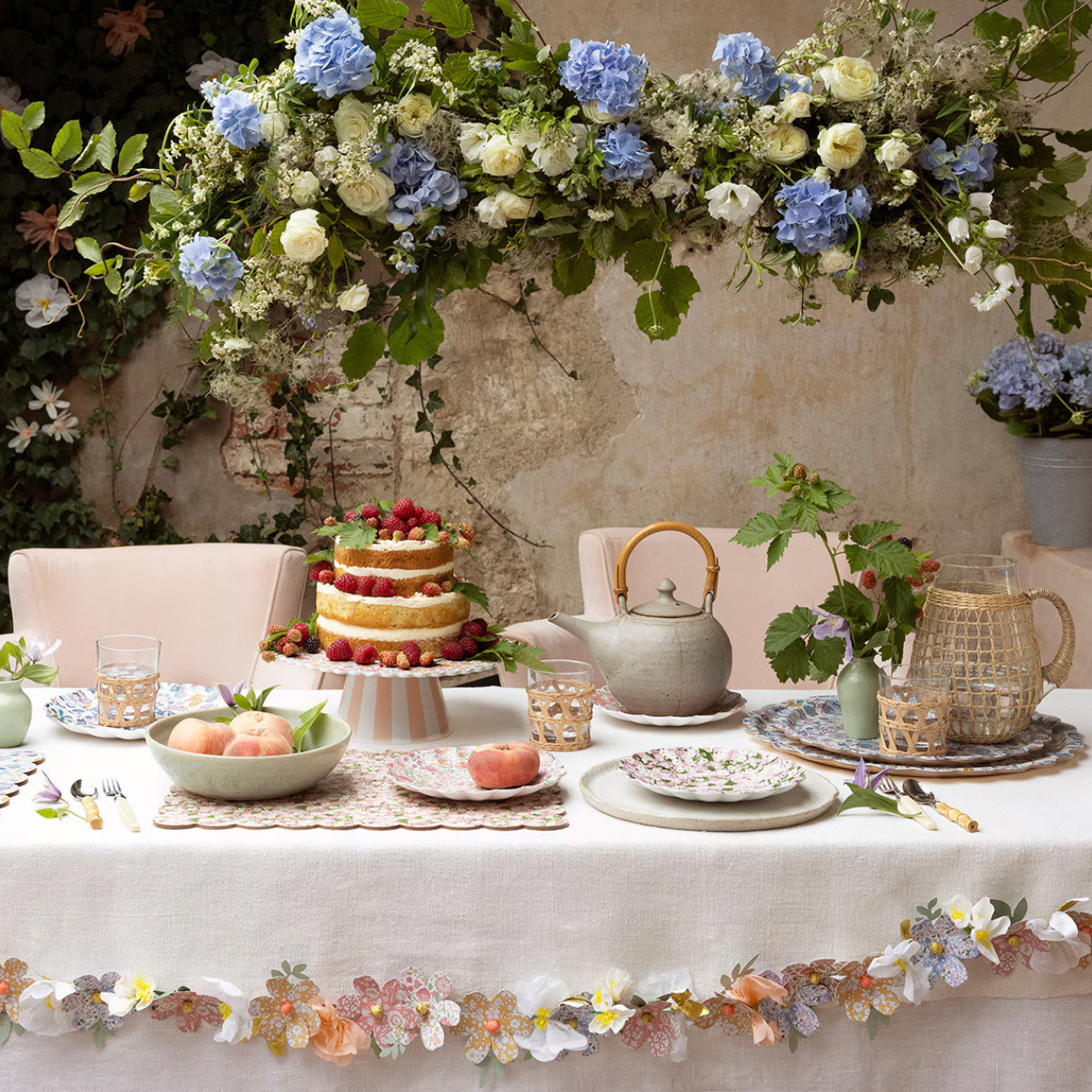Decorative table setting with flowers, cake, and teapots against a stone wall.