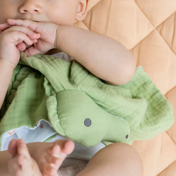 Baby wearing a green bib with a pattern of small animals, sitting on a wooden floor.