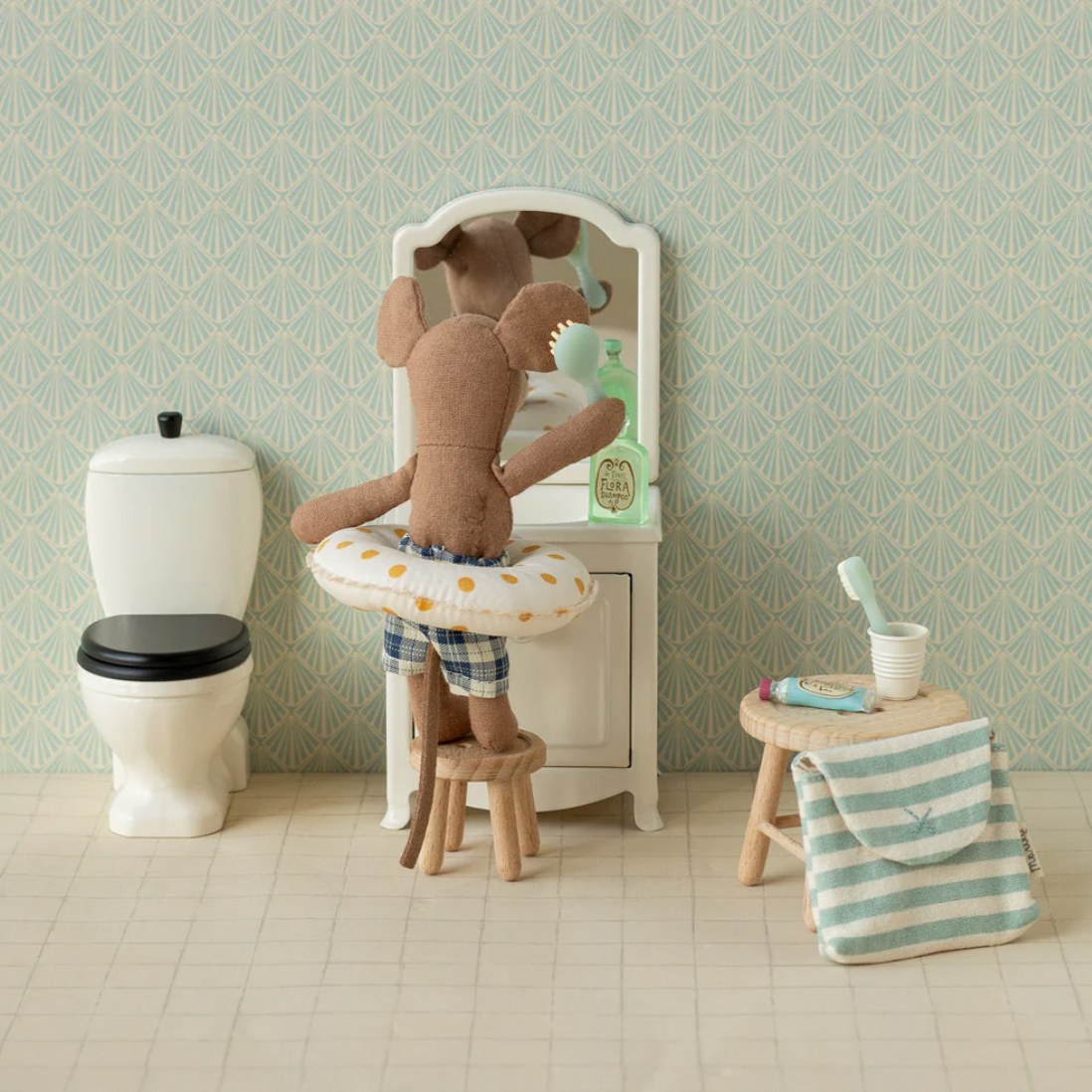 Miniature bathroom setup with a teddy bear on a toilet, sink, and stool against a patterned wall.