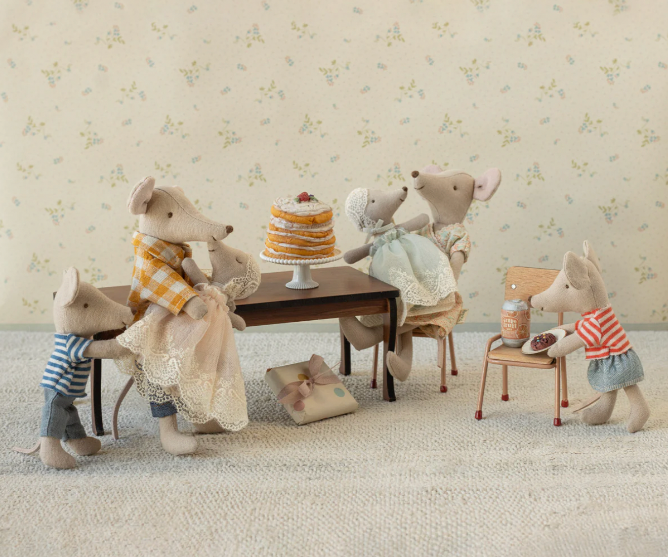 Stuffed animal mice gathered around a table with a cake in a room with floral wallpaper.
