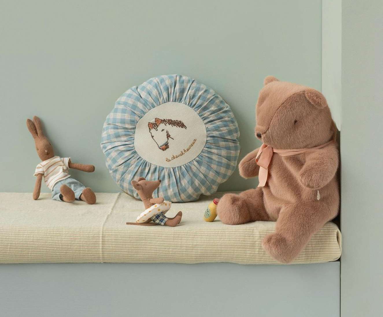 Children's toys including a teddy bear and dolls on a cushion against a light green wall.