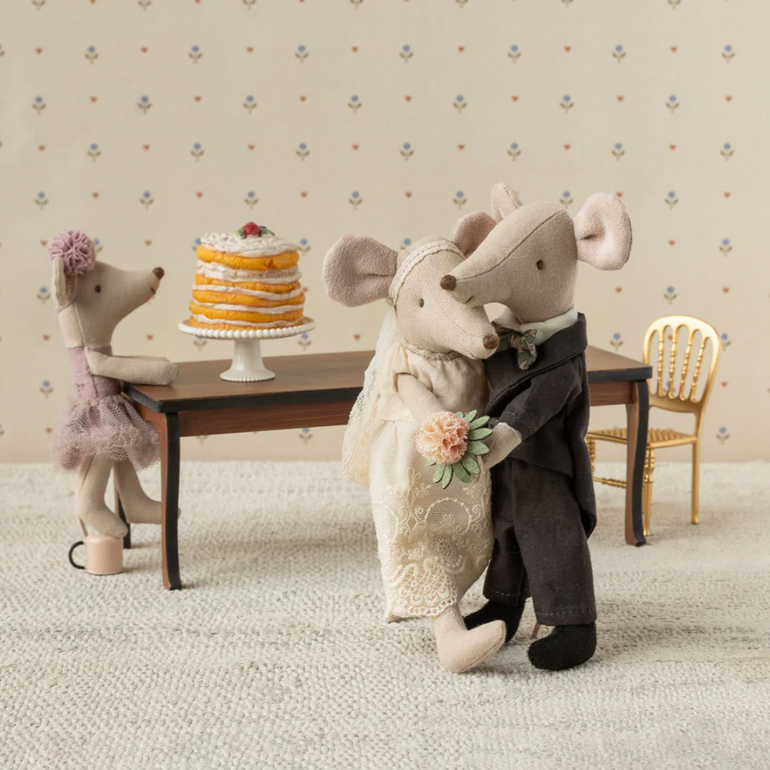 Two stuffed mouse figures in wedding attire with a cake on a table, set against a neutral wallpaper background.