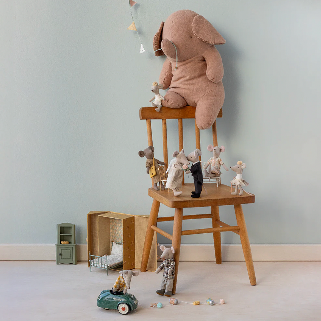 Stuffed elephant toy on a wooden stool with small figurines against a light gray wall.