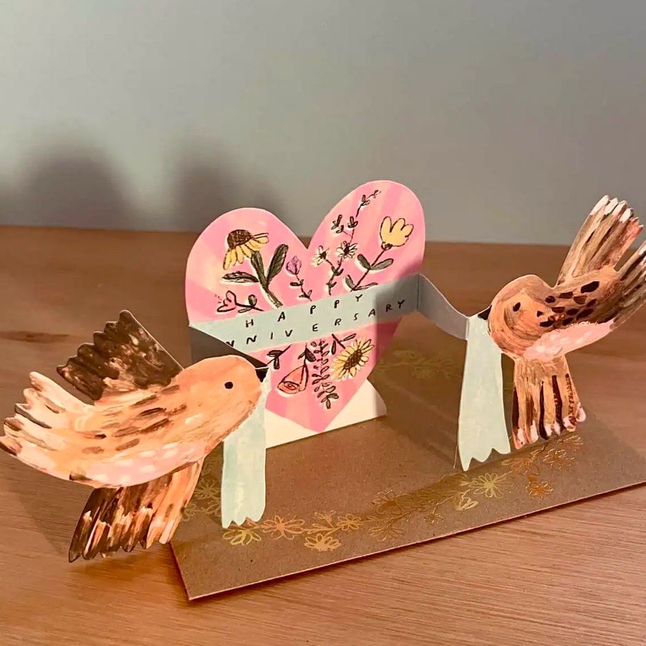 3D paper craft of two birds with a heart-shaped background on a wooden surface.
