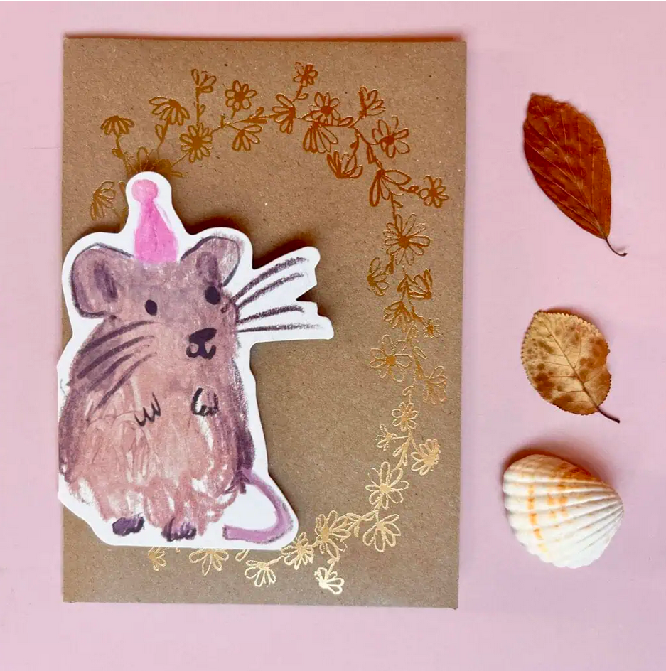 card of a mouse with a party hat on a brown envelope with floral patterns, surrounded by leaves and a shell on a pink background.