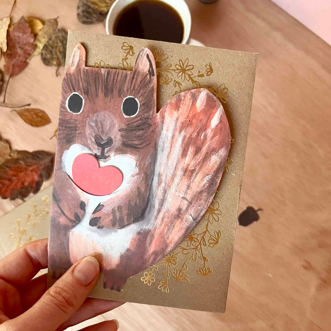 Card with a squirrel holding a heart on a wooden surface with a cup of coffee.