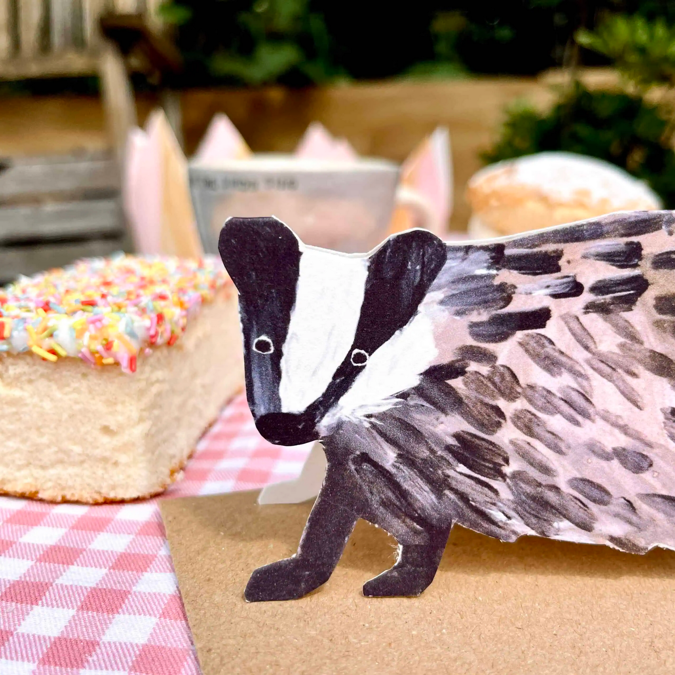 Cardboard card of a badger on a checkered tablecloth with a cake in the background