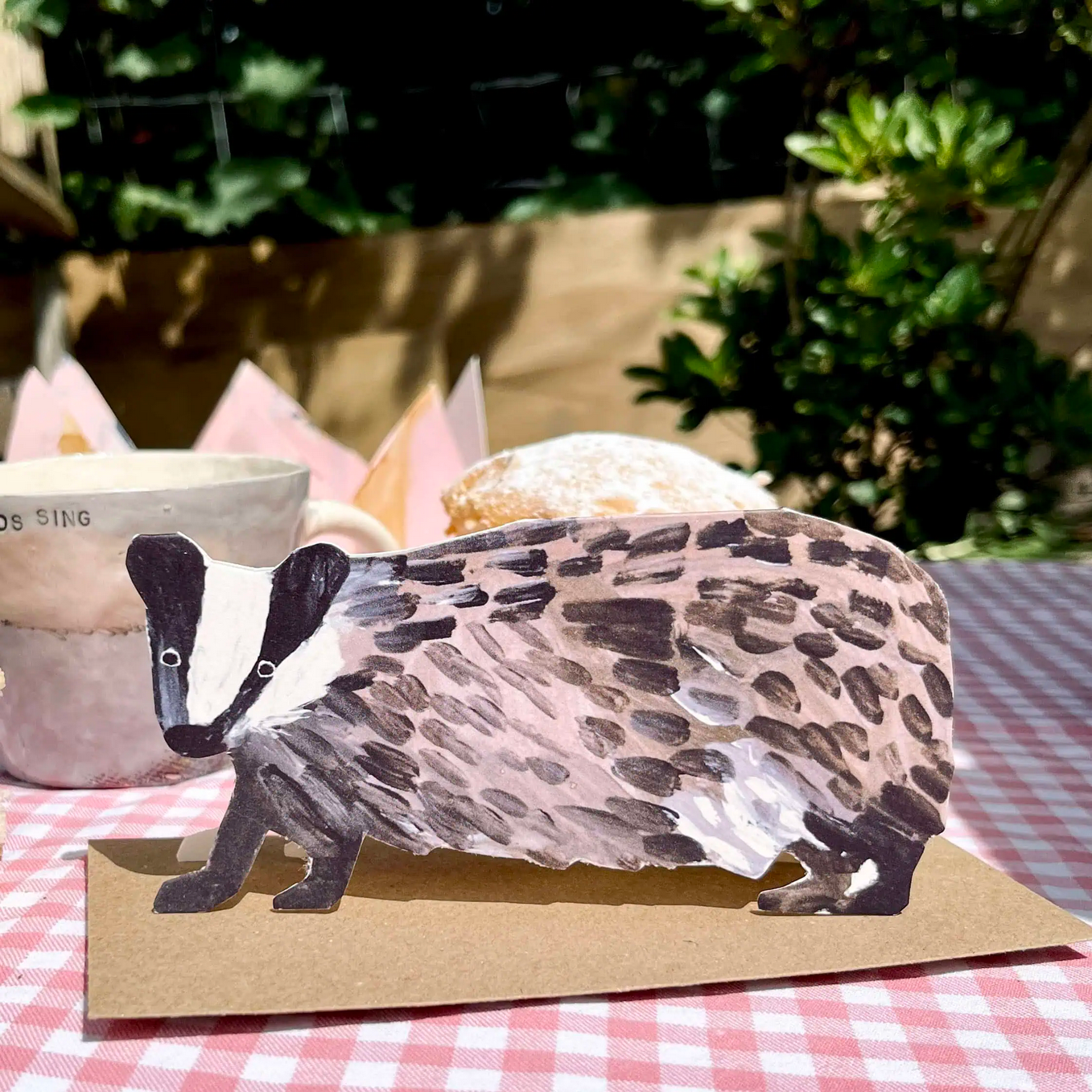 Cardboard card of a badger on a checkered tablecloth with plants in the background