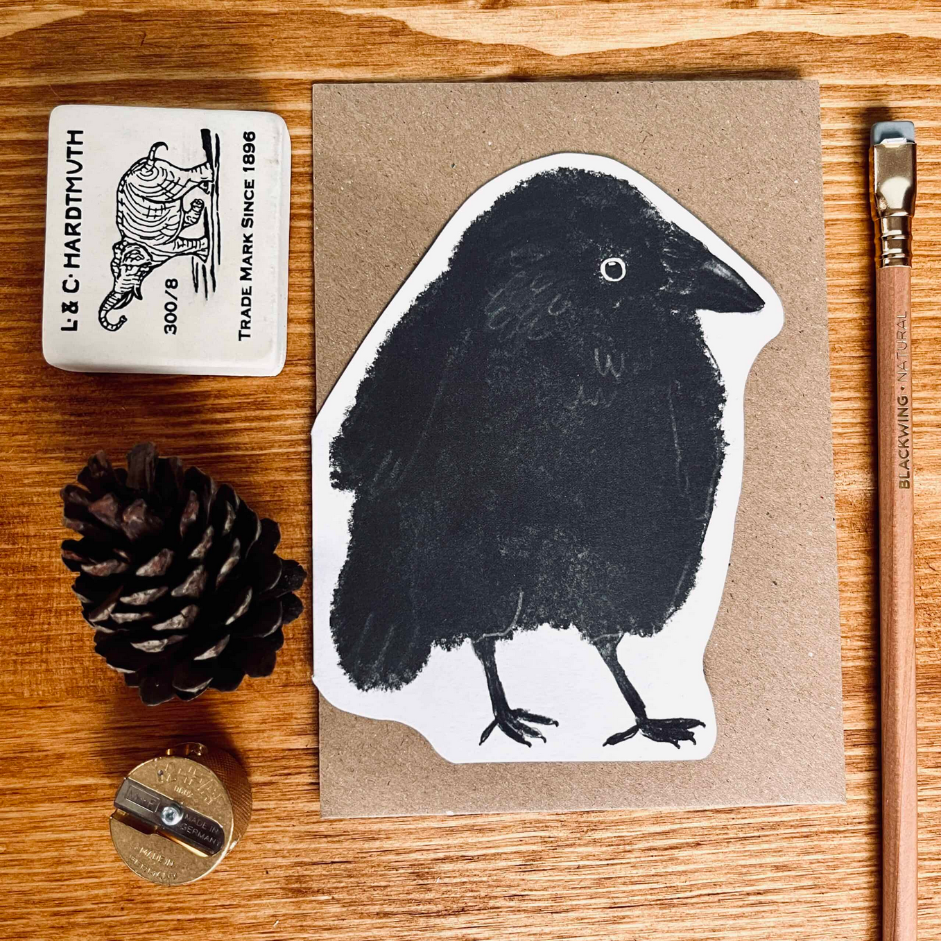 Black and white bird illustration on a brown card with stationery items on a wooden surface