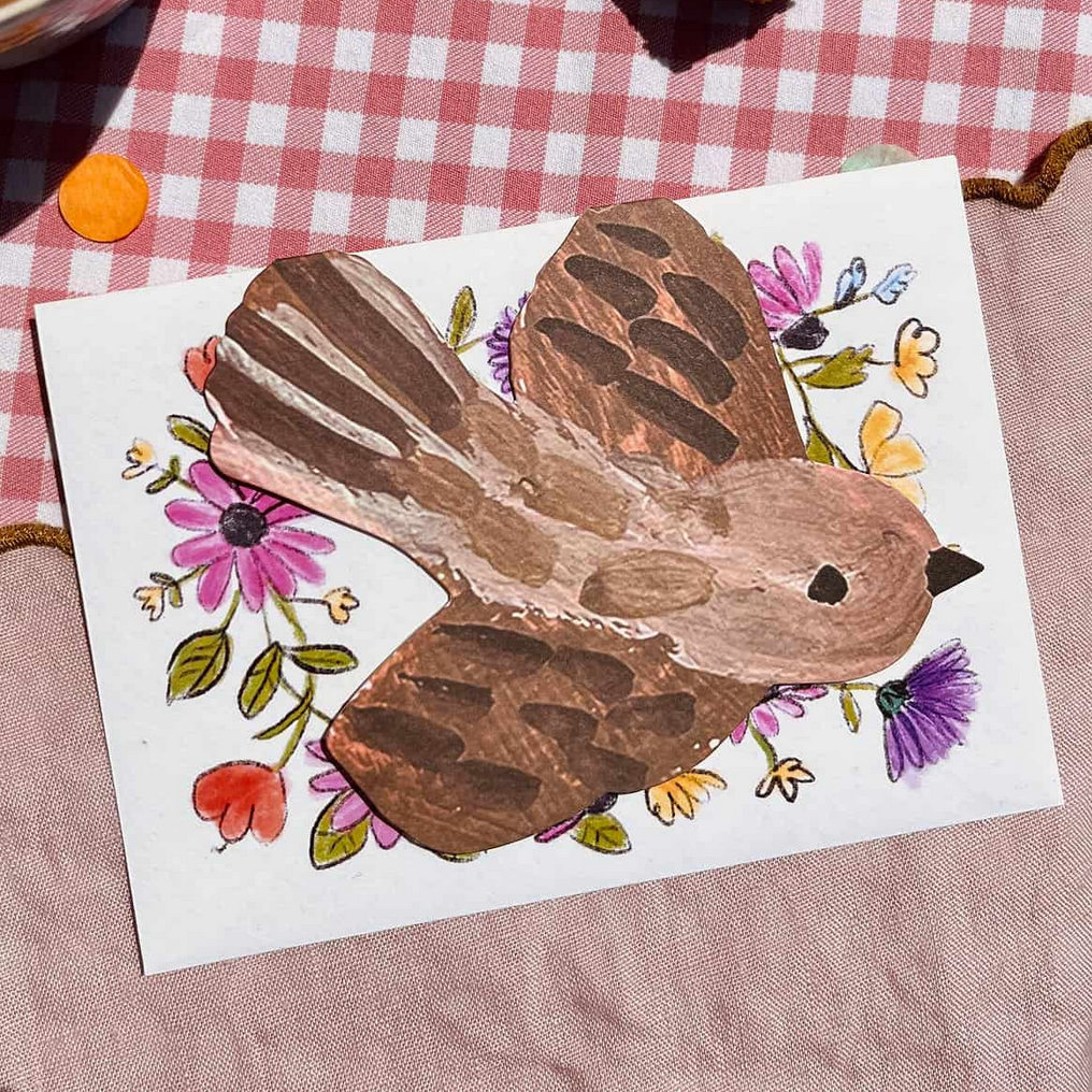 Wooden bird decoration on a card with flowers, next to a cup of coffee and cake on a checkered tablecloth.