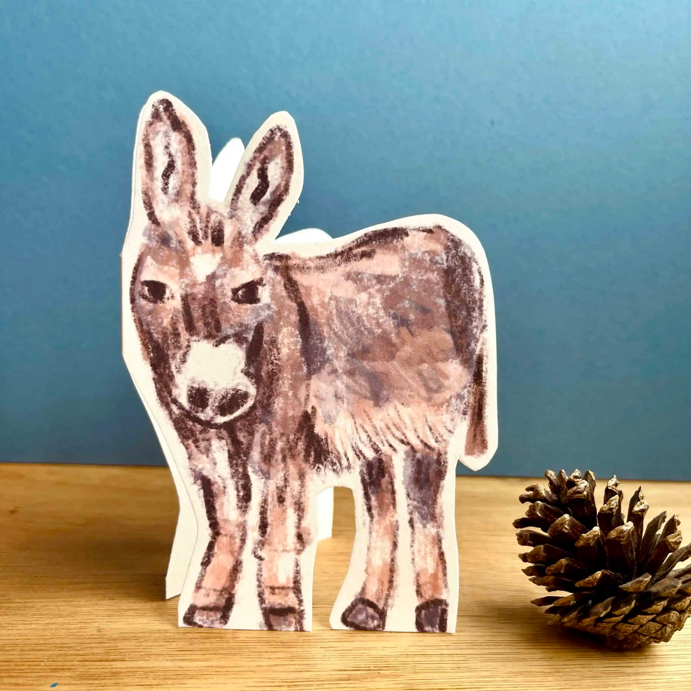 Wooden donkey card on a wooden surface with a pine cone against a blue background