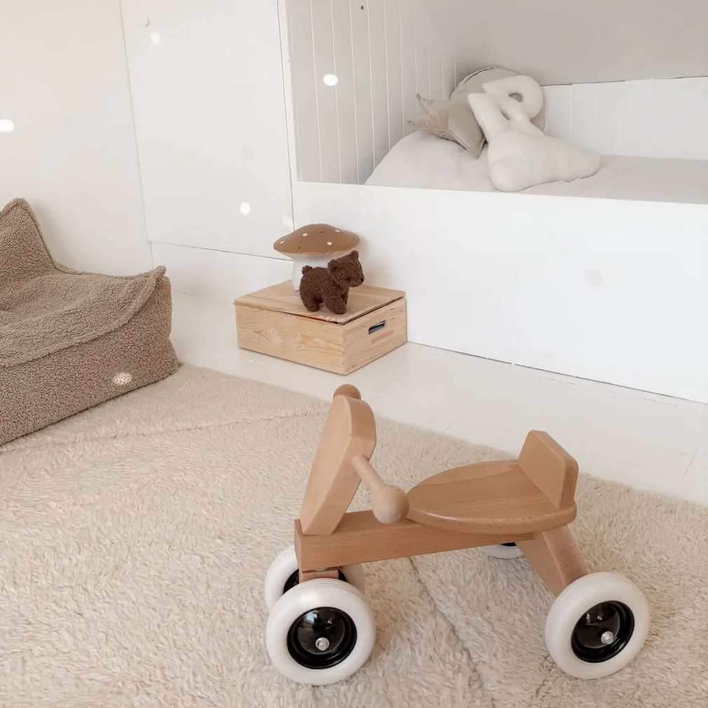 Wooden toy car on a carpeted floor in a child's room with a bed and toys in the background.