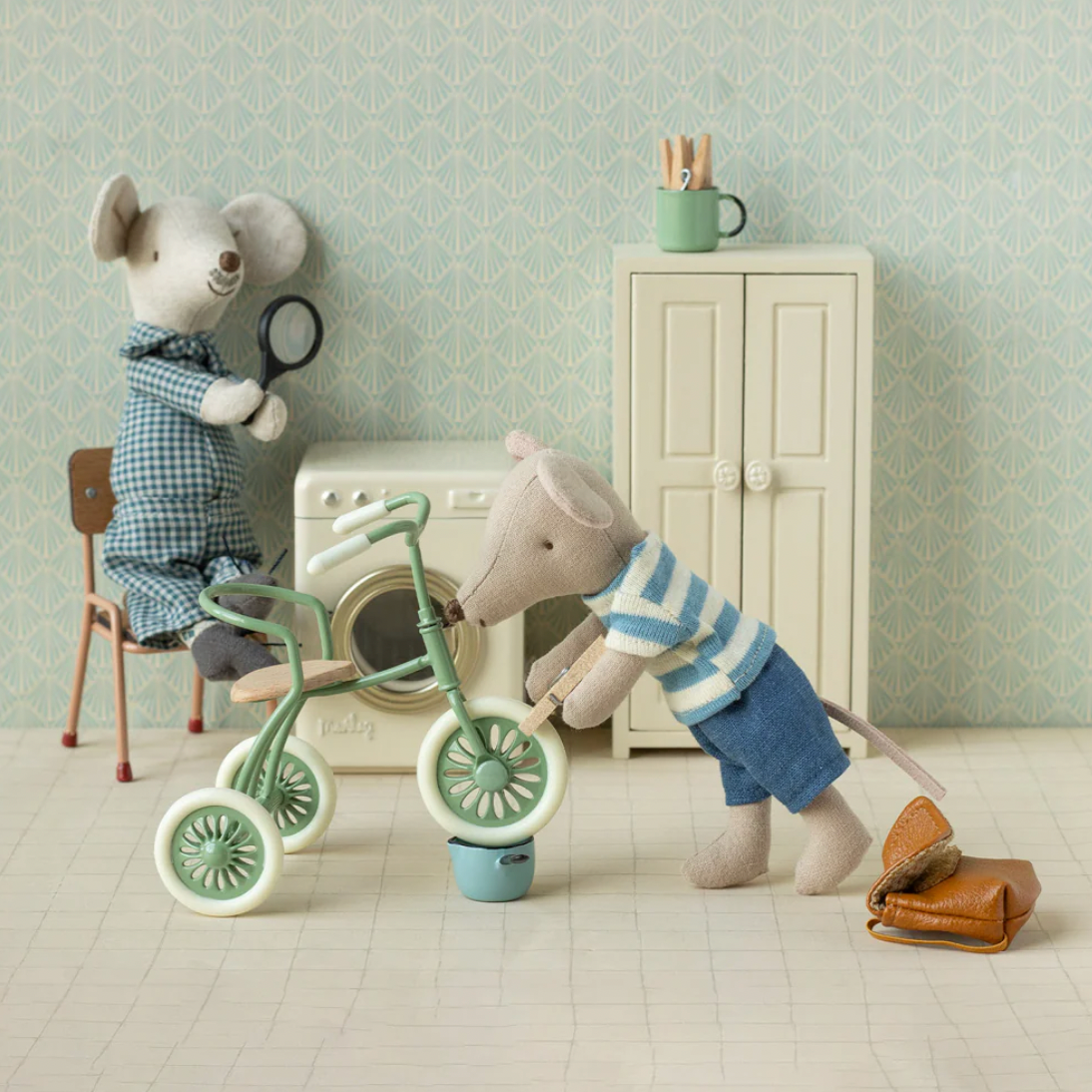 Two toy mice with a tricycle in a room setting, featuring a washing machine and cabinet.